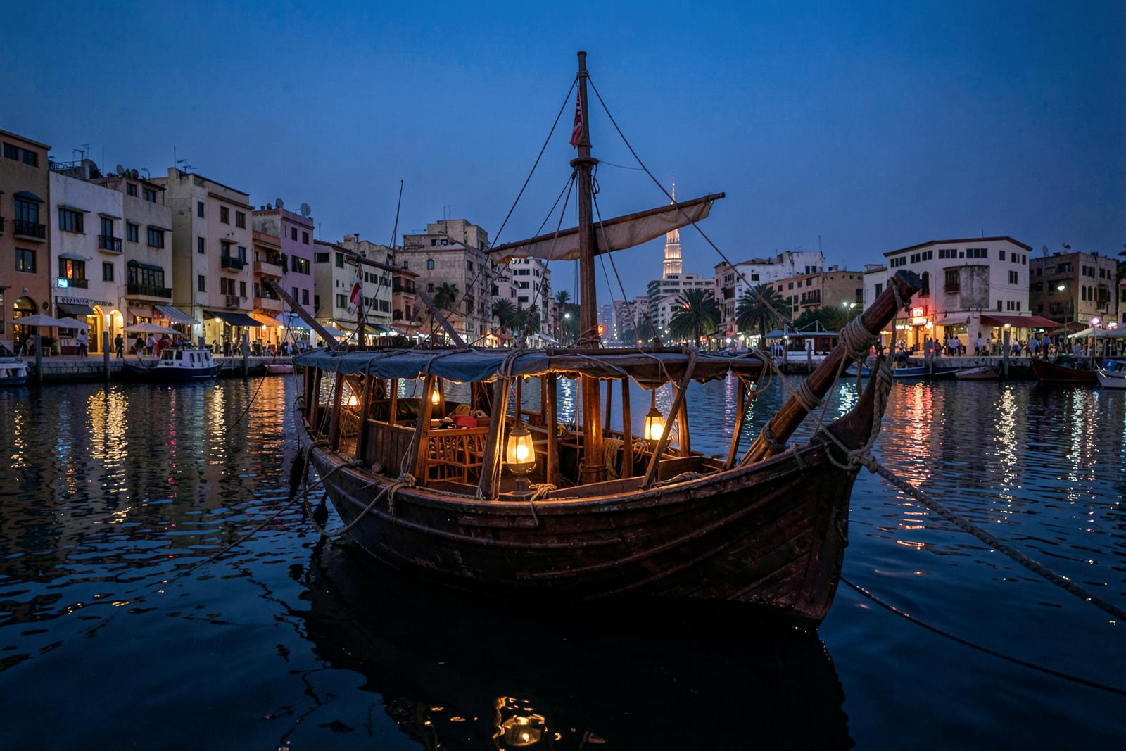Illustrative scene: a wooden dhow with lit lanterns moored on a creek in front of low heritage buildings at dusk.